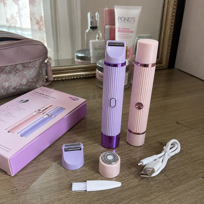 A pink and purple electric facial and body razor set displayed on a wooden table with its pink retail box, charging cable, cleaning brush, and interchangeable heads. A mirror in the background shows other beauty products, including a tube of Pond's.